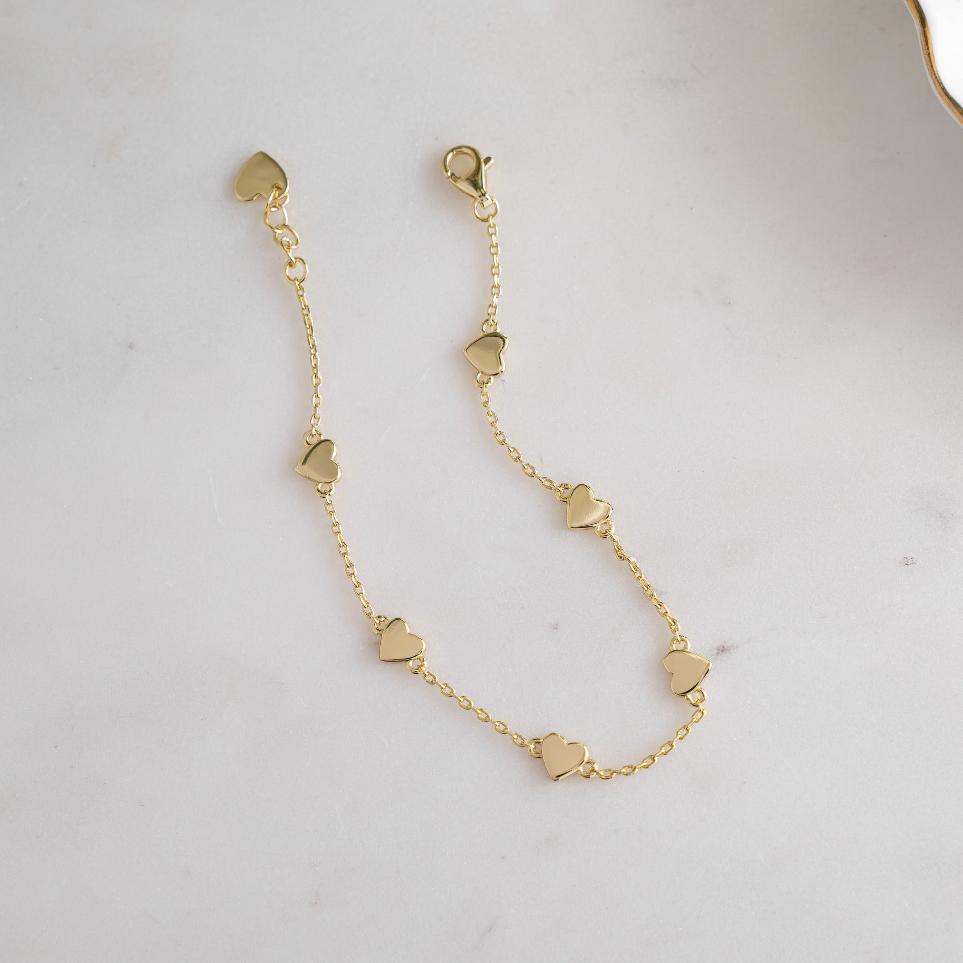 Gold charm bracelet laid flat, showcasing its dainty heart charms, fine chain and clasp. 