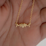 Model's hand showcasing dainty necklace in gold with 'MOM' heart pendant. 