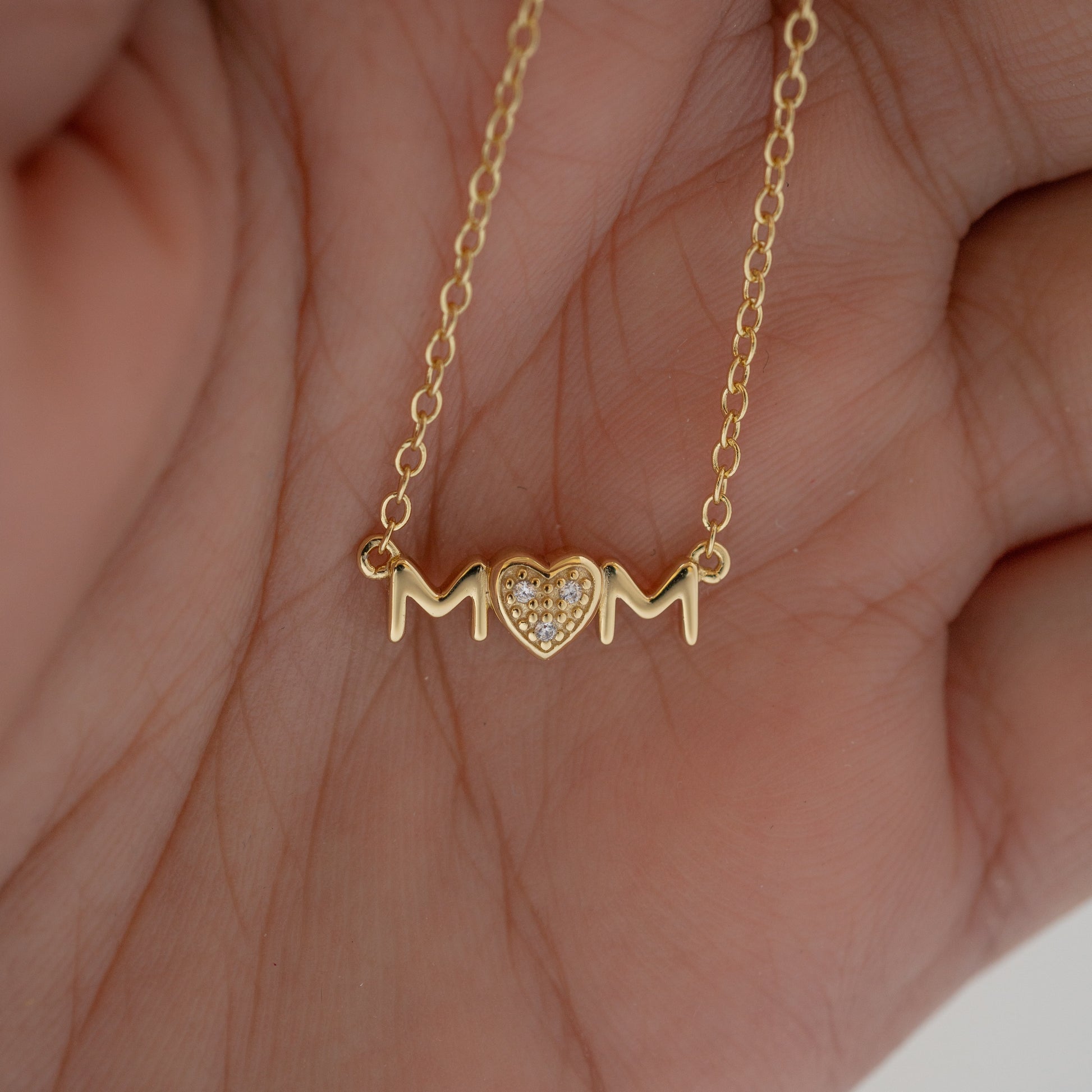 Model's hand showcasing dainty necklace in gold with 'MOM' heart pendant. 