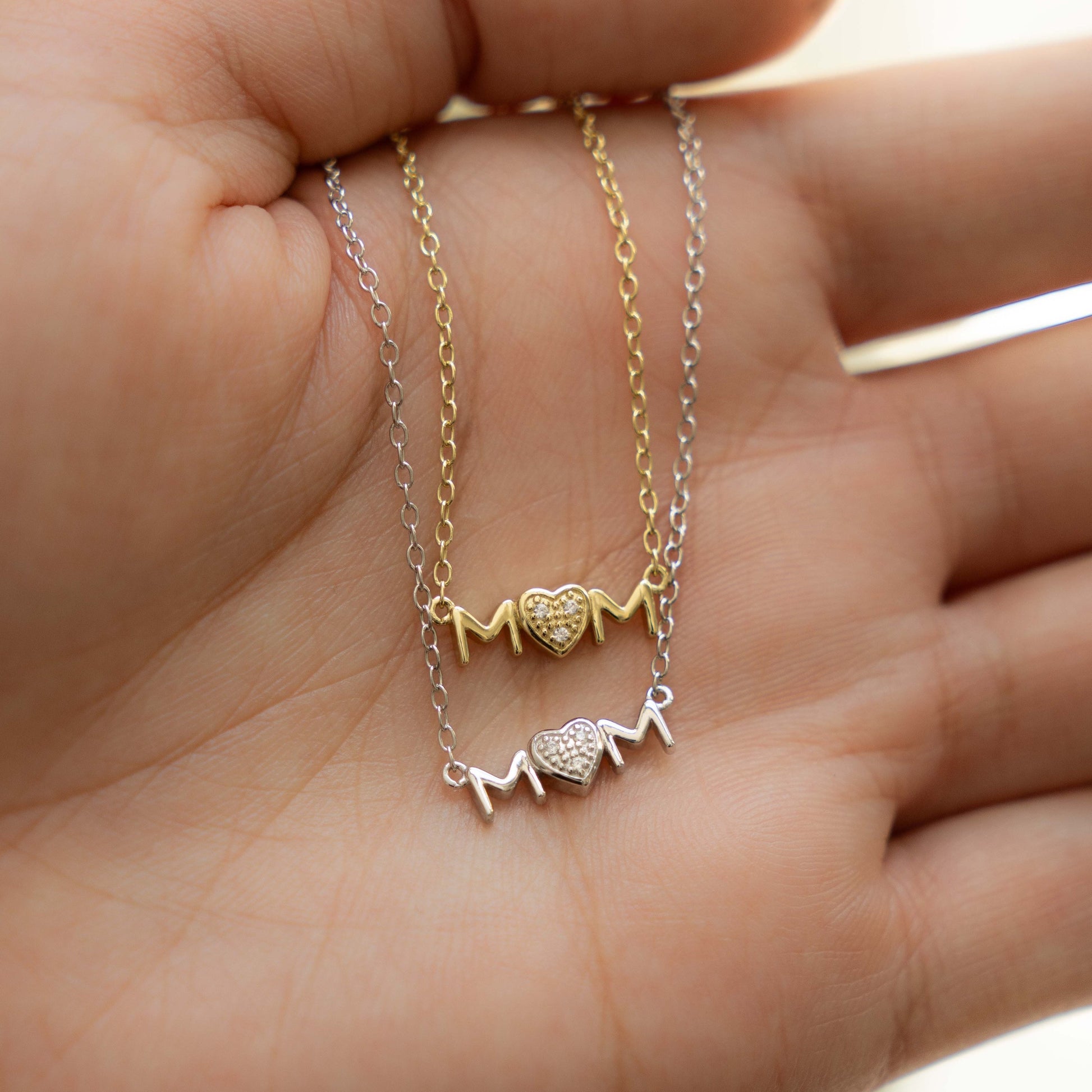 Model's hand showcasing dainty necklaces, one gold pendant necklace and one silver pendant necklace, both featuring a "MOM" heart pendant