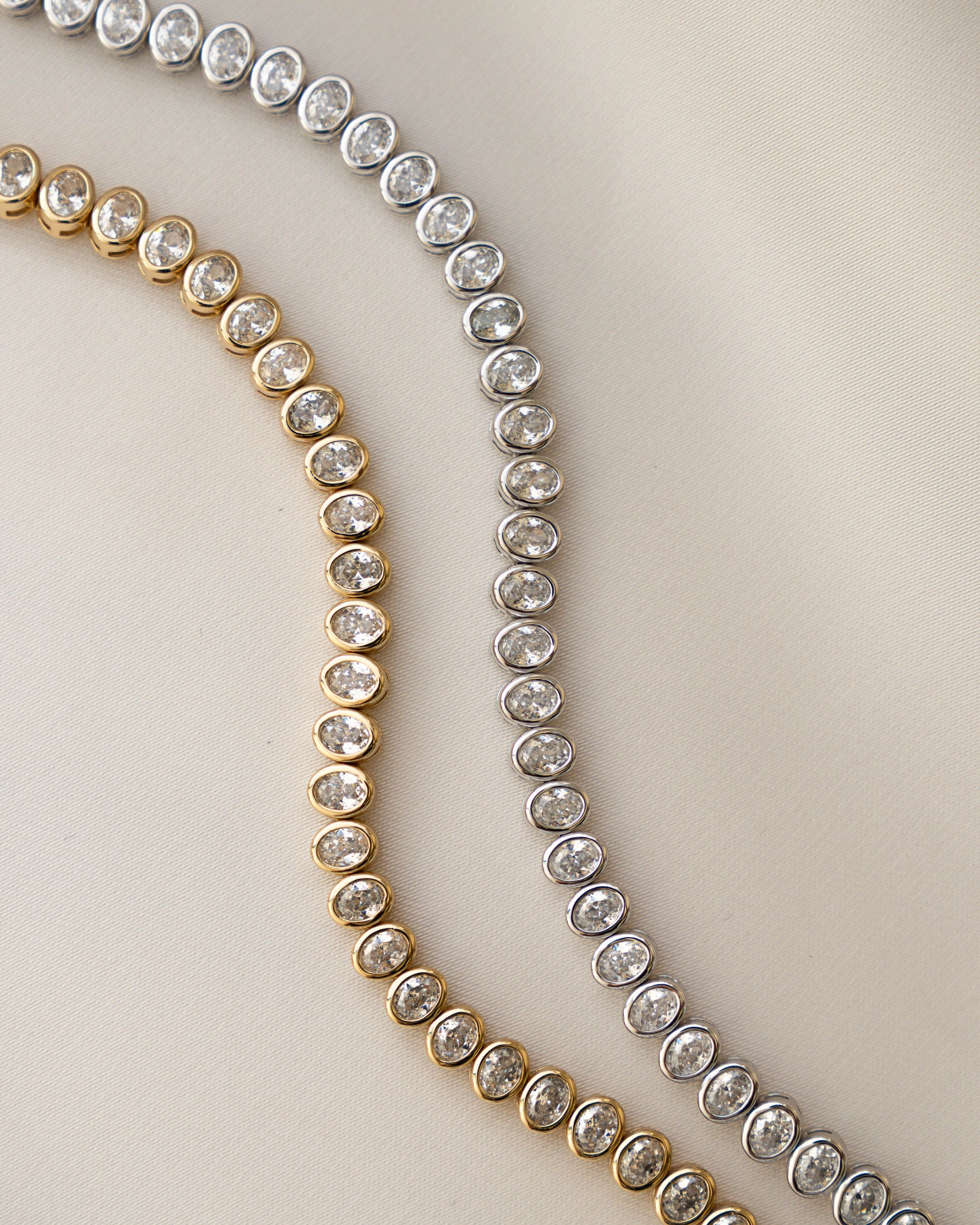 closeup image of two stackable diamond tennis bracelets featuring a silver oval bezel set diamond tennis bracelet and a gold 14k plated oval diamond bracelet laying on a beige surface