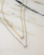 gold pendant necklace with paperclip chain laying next to a sterling silver baguette necklace