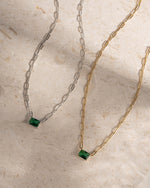 Two necklaces, one gold paperclip necklace and one silver paperclip necklace, with emerald pendant on a beige surface.
