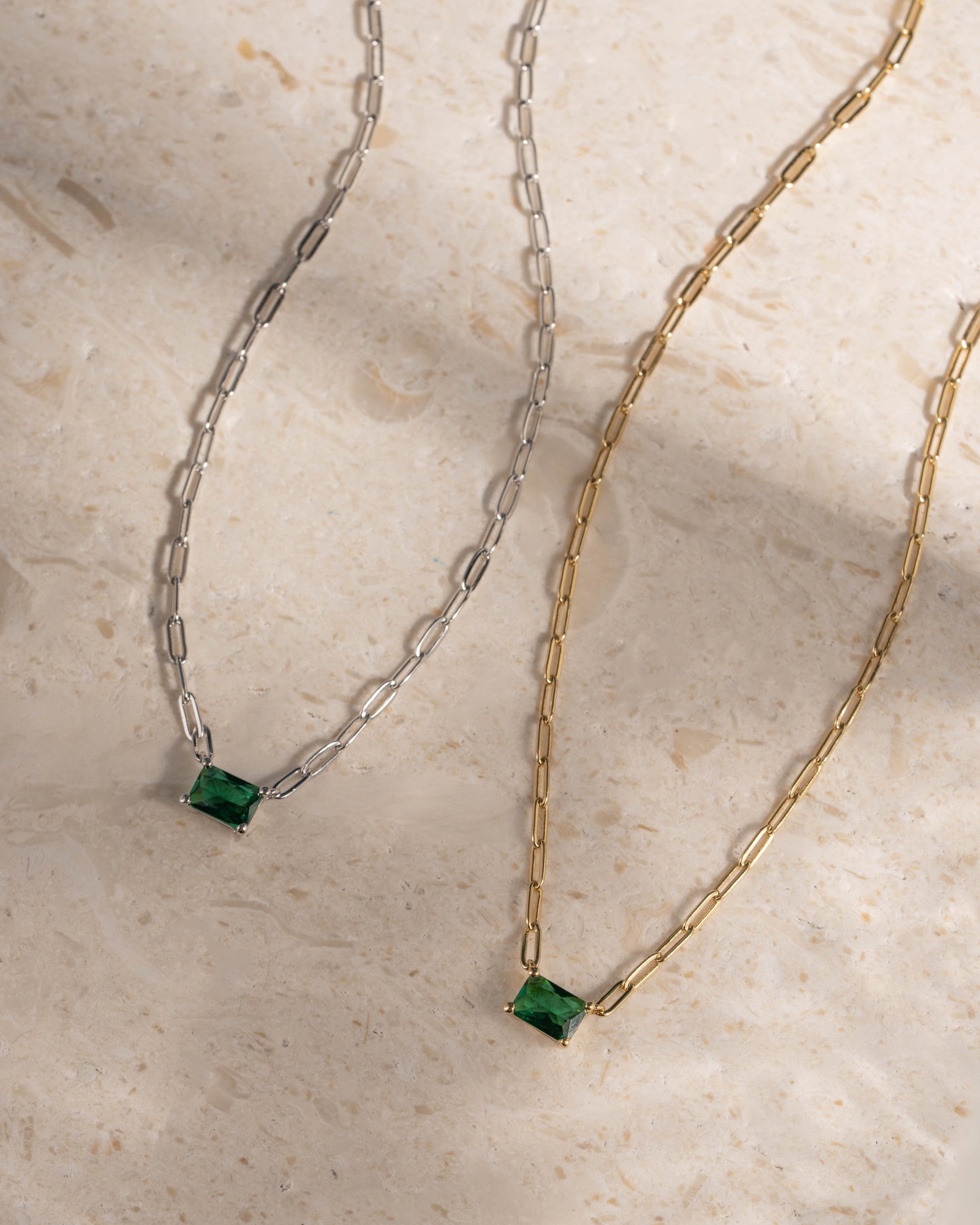 Two necklaces, one gold paperclip necklace and one silver paperclip necklace, with emerald pendant on a beige surface.