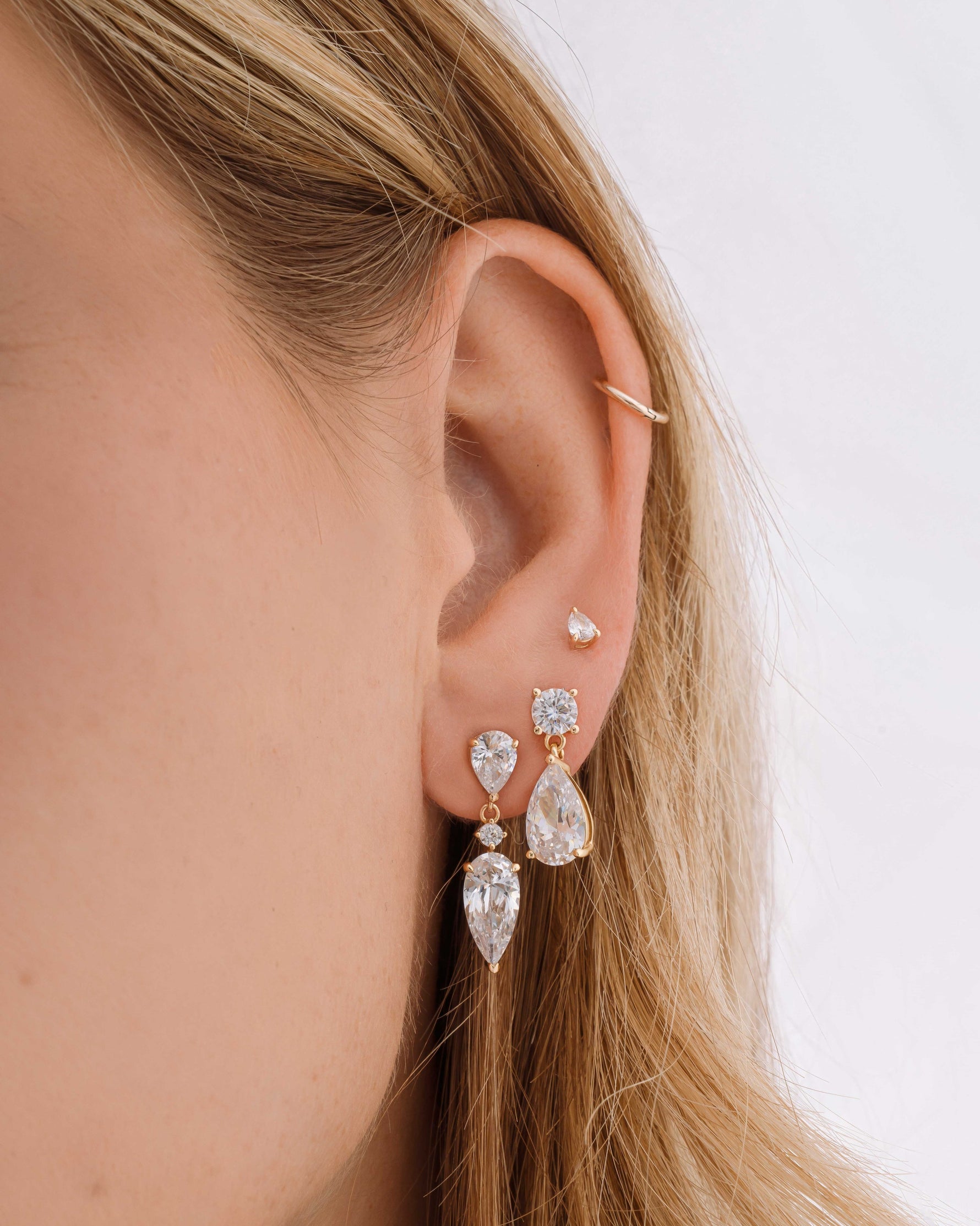 Gold diamond earrings on model featuring drop earrings with a pear cut, followed by diamond charm earrings and a pear cut diamond stud earring in models third hole