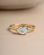 Gold bridesmaid jewelry featuring a gold ring with a pear-shaped diamond on a beige surface