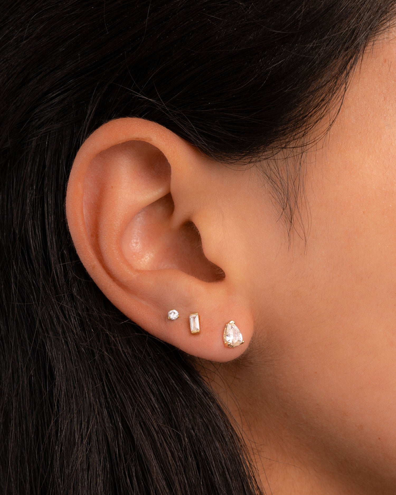 Model wearing gold pear stud earrings, a tiny baguette stud earrings, and a solitaire stud earrings