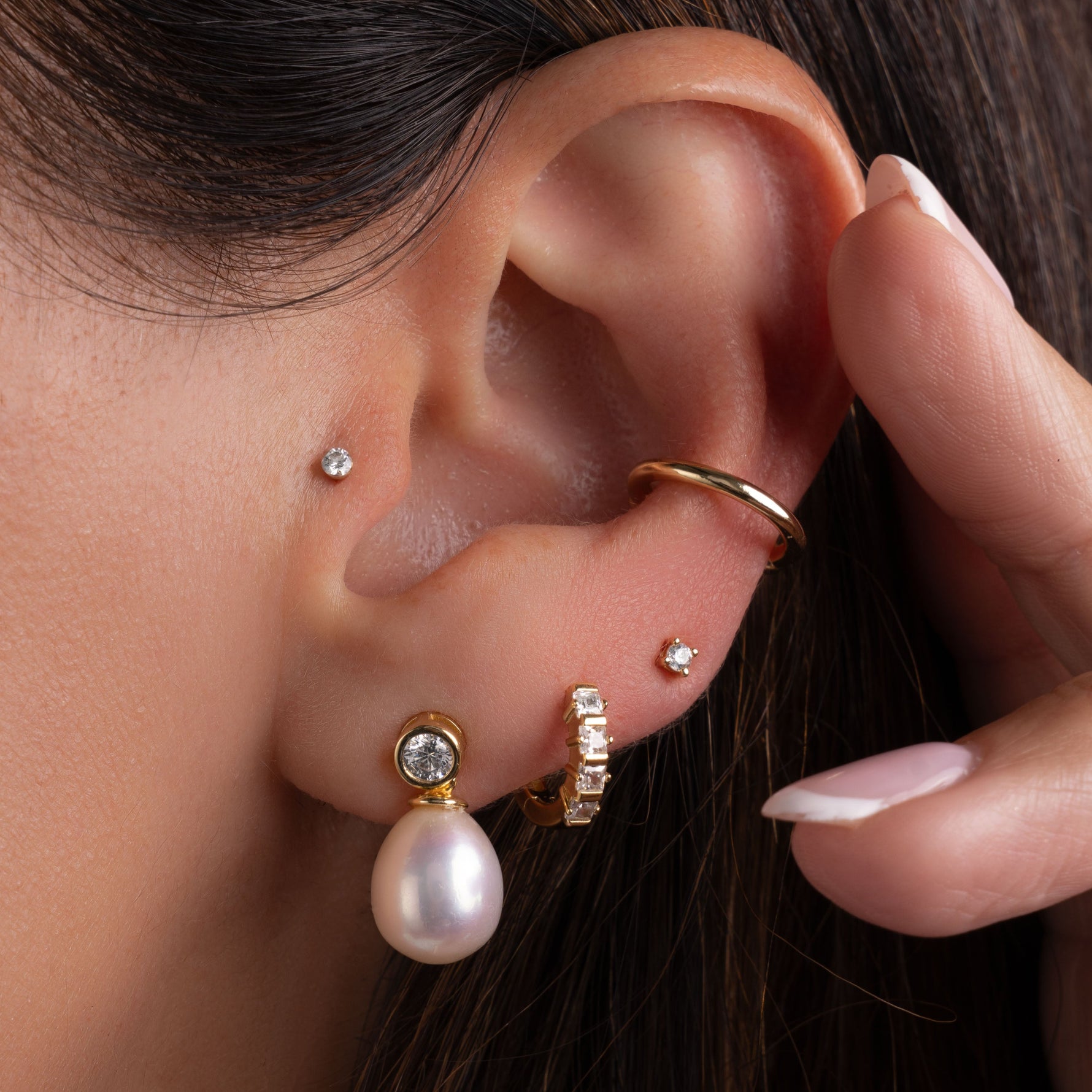 model wearing a gold ear stack featuring a freshwater pearl dangling earring, a huggie hoop earring, a simple cz stud earring and an ear cuff