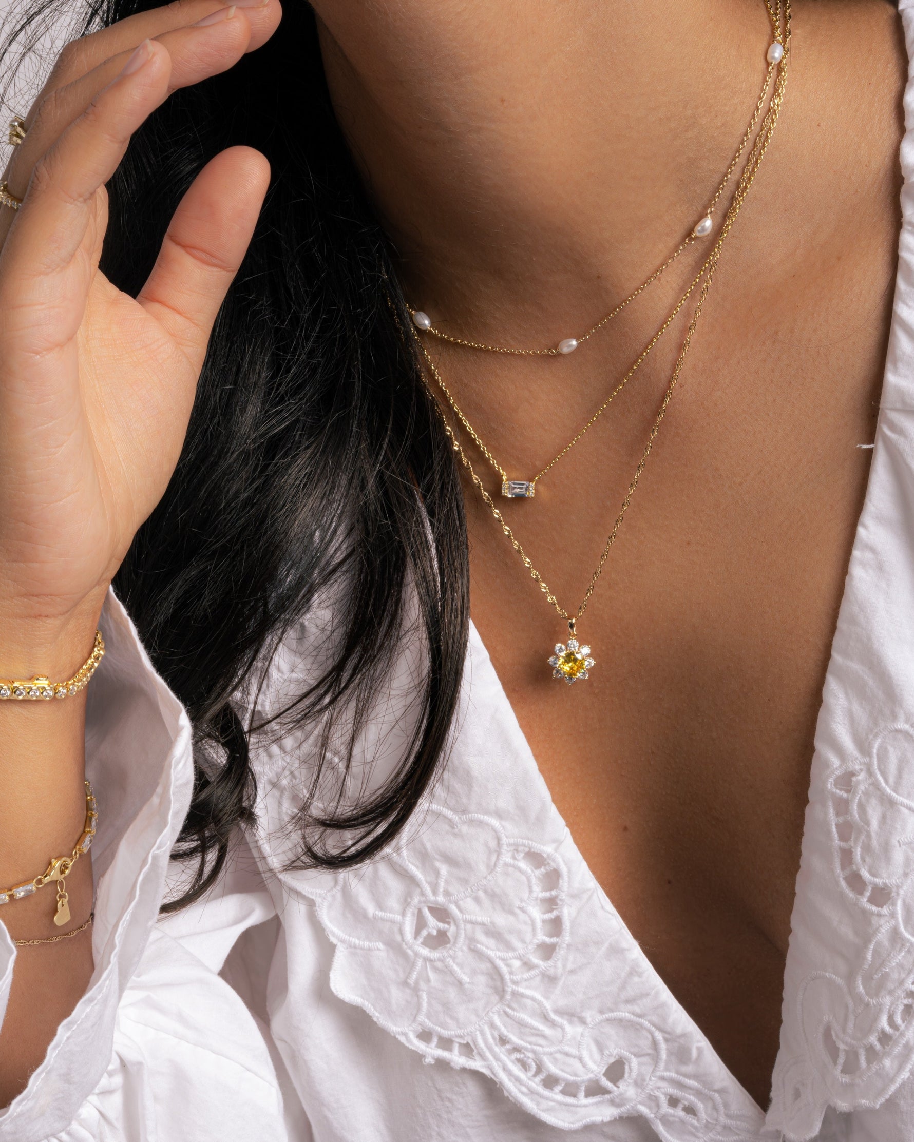 Model wearing a delicate gold necklaces with a white lace top featuring a beaded pearl necklace, a baguette diamond necklace, and a sunflower pendant necklace