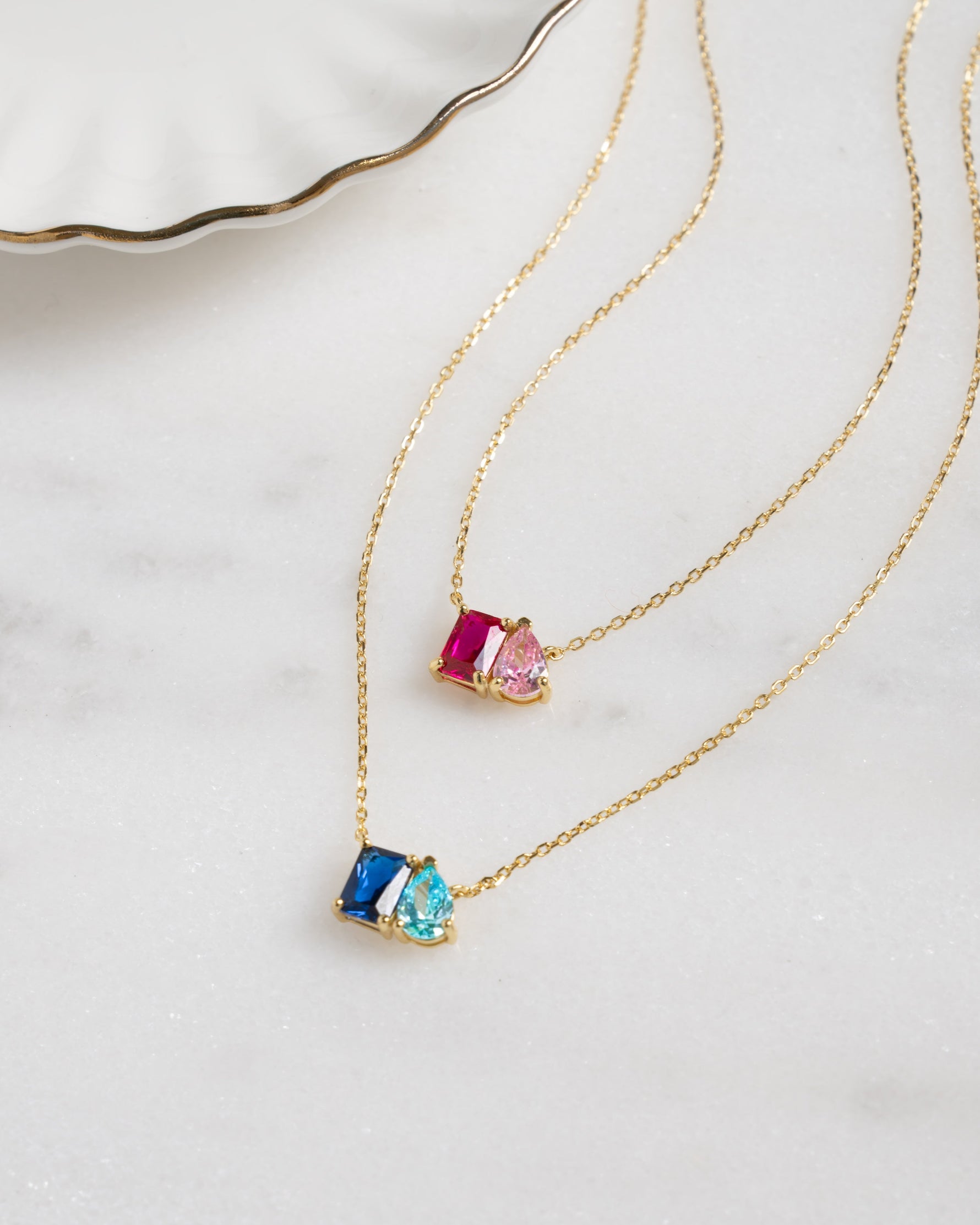 fatlay image of toi et moi birthstone necklaces with custom birthstone gemstone, one gold necklace features a ruby and rose birthstone, while the other gold necklace features a sapphire and aqua birthstone