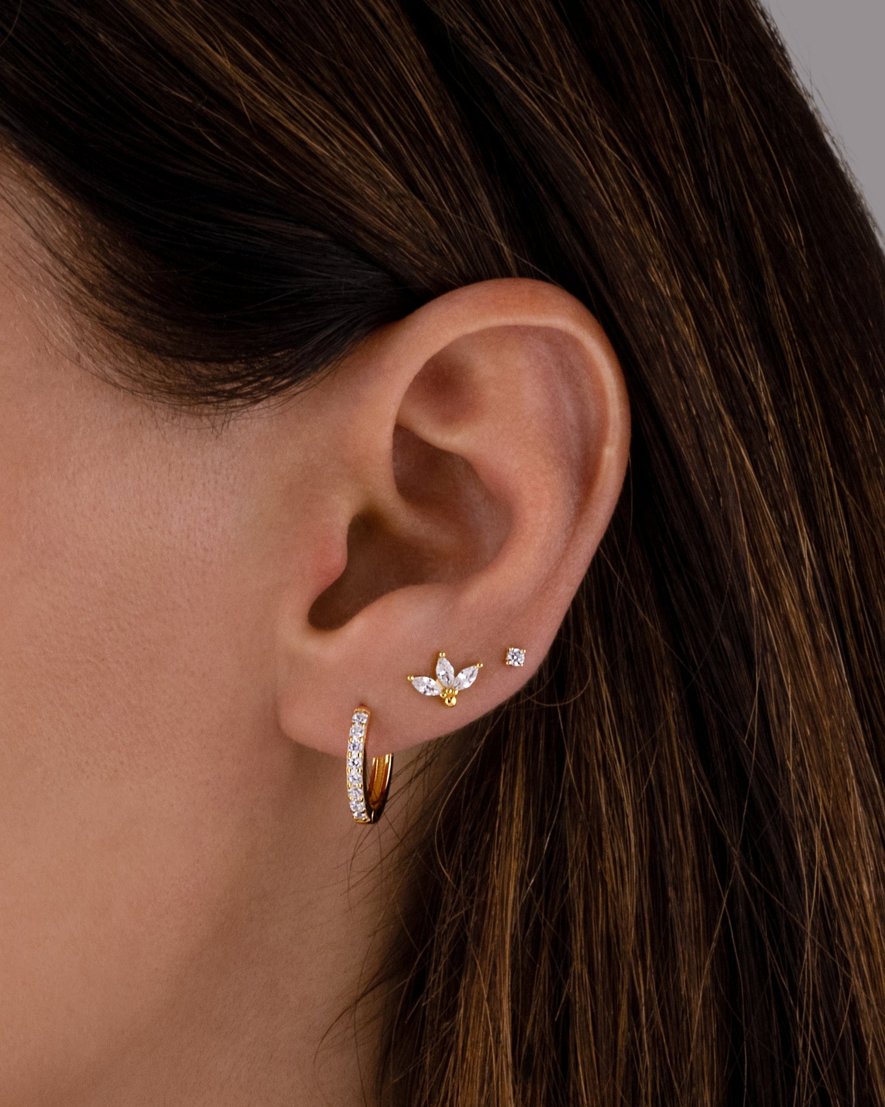 closeup image of model wearing cubic zirconia studded hoop earrings, dainty gold petal stud earrings, and a solitaire stud earrings