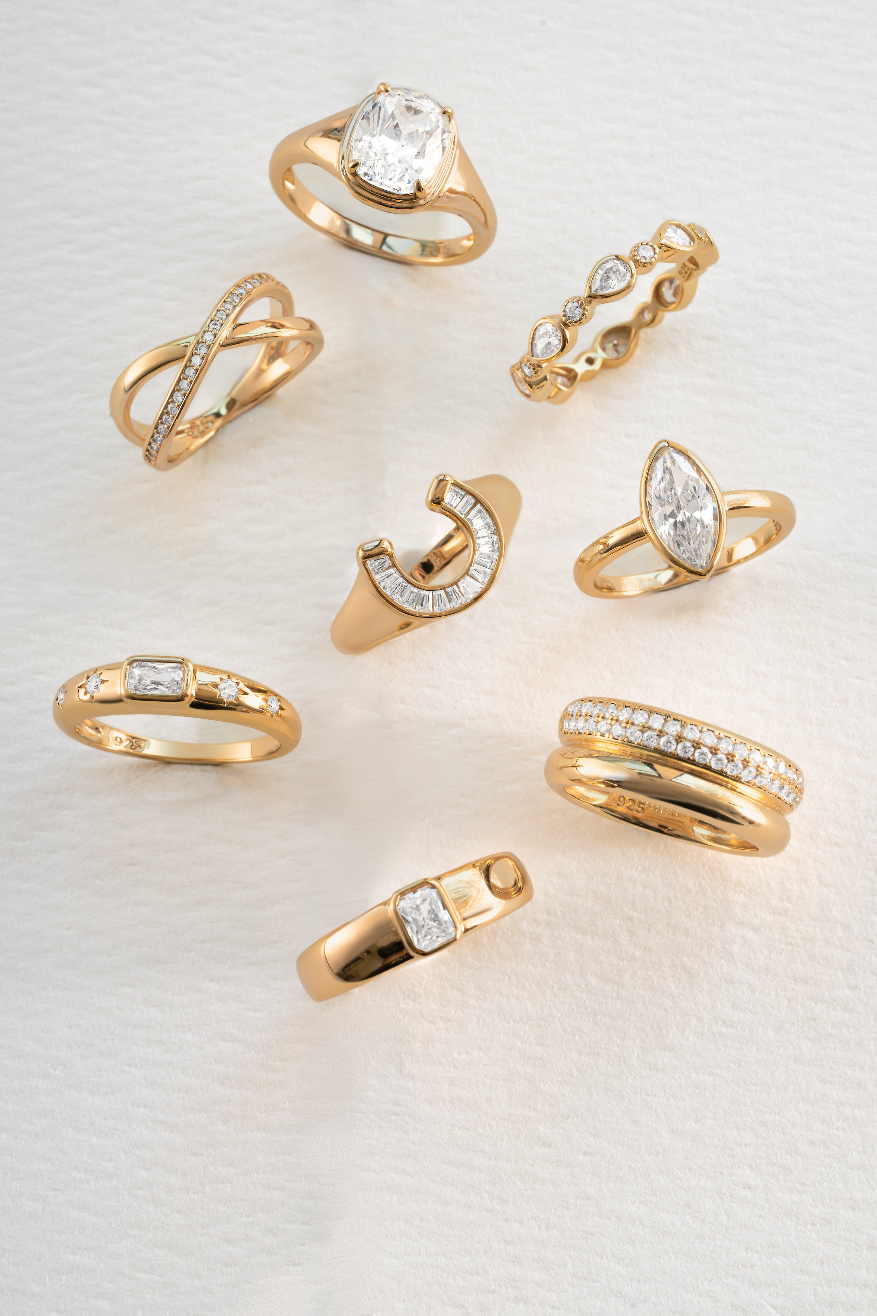 A set of nine yellow gold rings with various designs, including pavé bands, a crescent moon with stones, and a ring with a large clear gemstone, arranged on a textured white background.