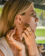 Woman adjusting earrings inside a car wearing gold stacking rings featuring a textured rope ring and multi stone ring, and gold earrings featuring a chunky hoop earring and simple gold hoop