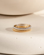 Gold ring with a textured band, and sterling silver stacking rope ring on a white ceramic surface