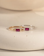 closeup image of two rings, one sterling silver ruby baguette ring and one gold ruby birthstone ring with a baguette center