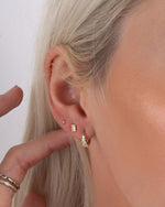 model touching her dainty gold earrings featuring a small hoop earring, a gold baguette stud earring, and a small gold ball stud earring
