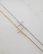 A gold cross bracelet with a sterling silver chain cross bracelet laying on a plain background