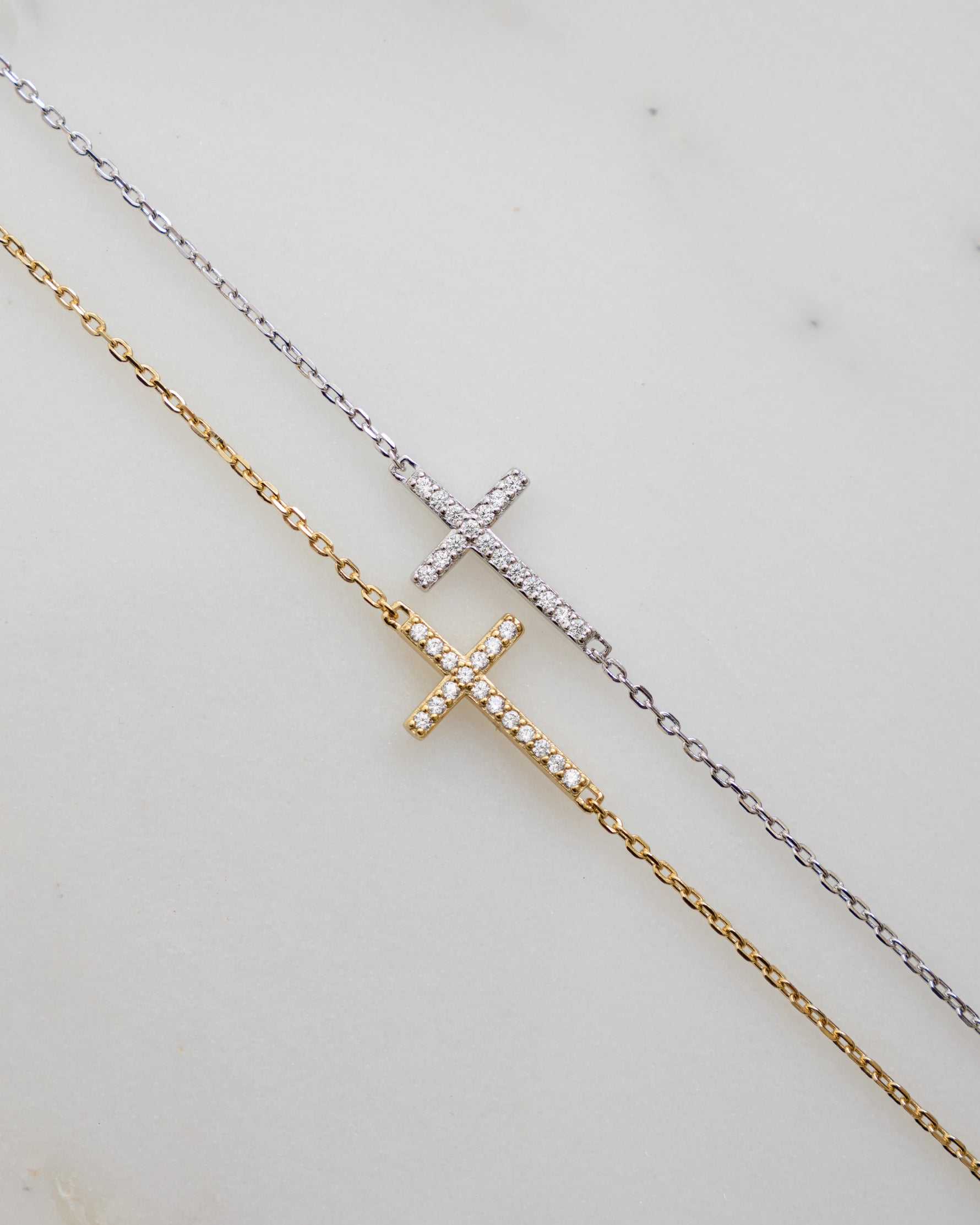 A gold cross bracelet with a sterling silver chain cross bracelet laying on a plain background