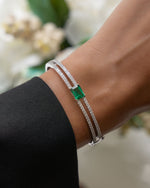 Close-up of a wrist wearing a silver statement bracelet with a emerald birthstone against a blurred floral background.