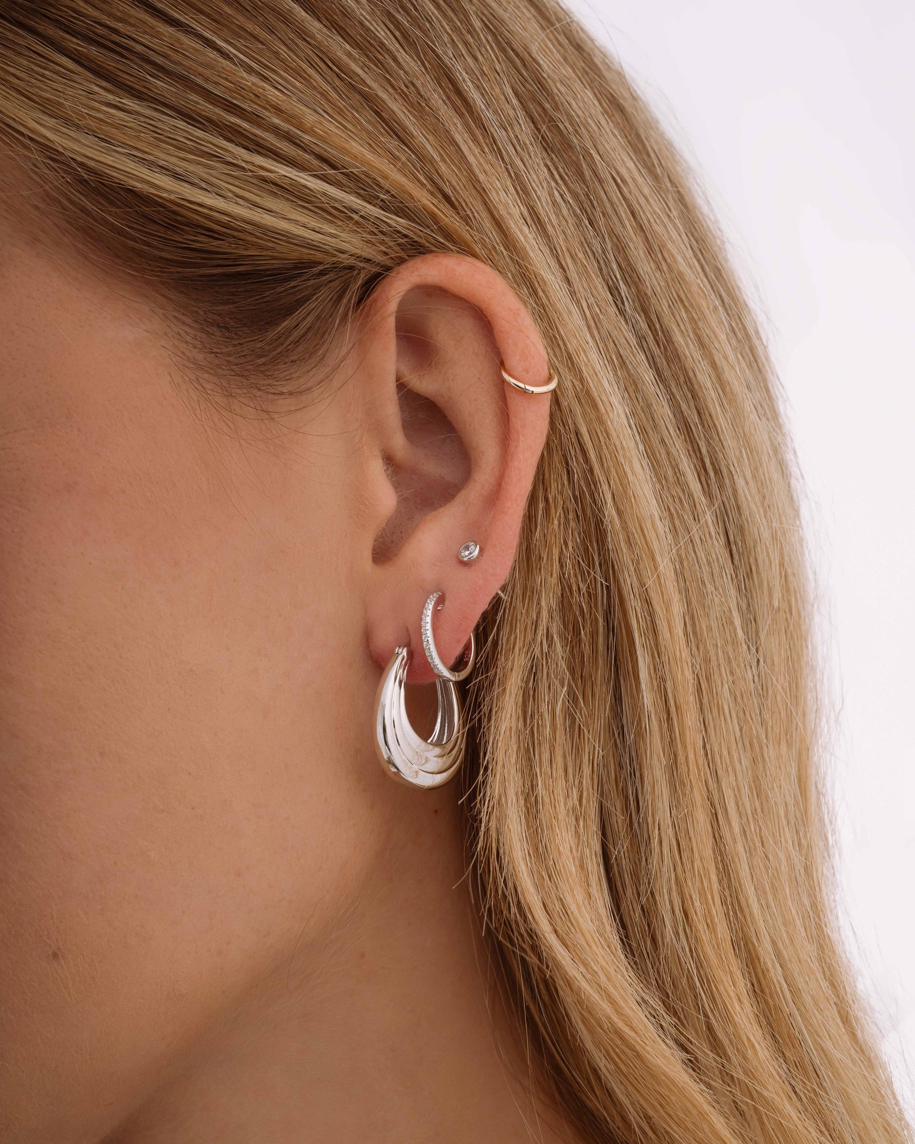 Closeup of statement earrings, featuring silver thick hoop earrings, diamond small hoop earrings, and silver stud earrings on model