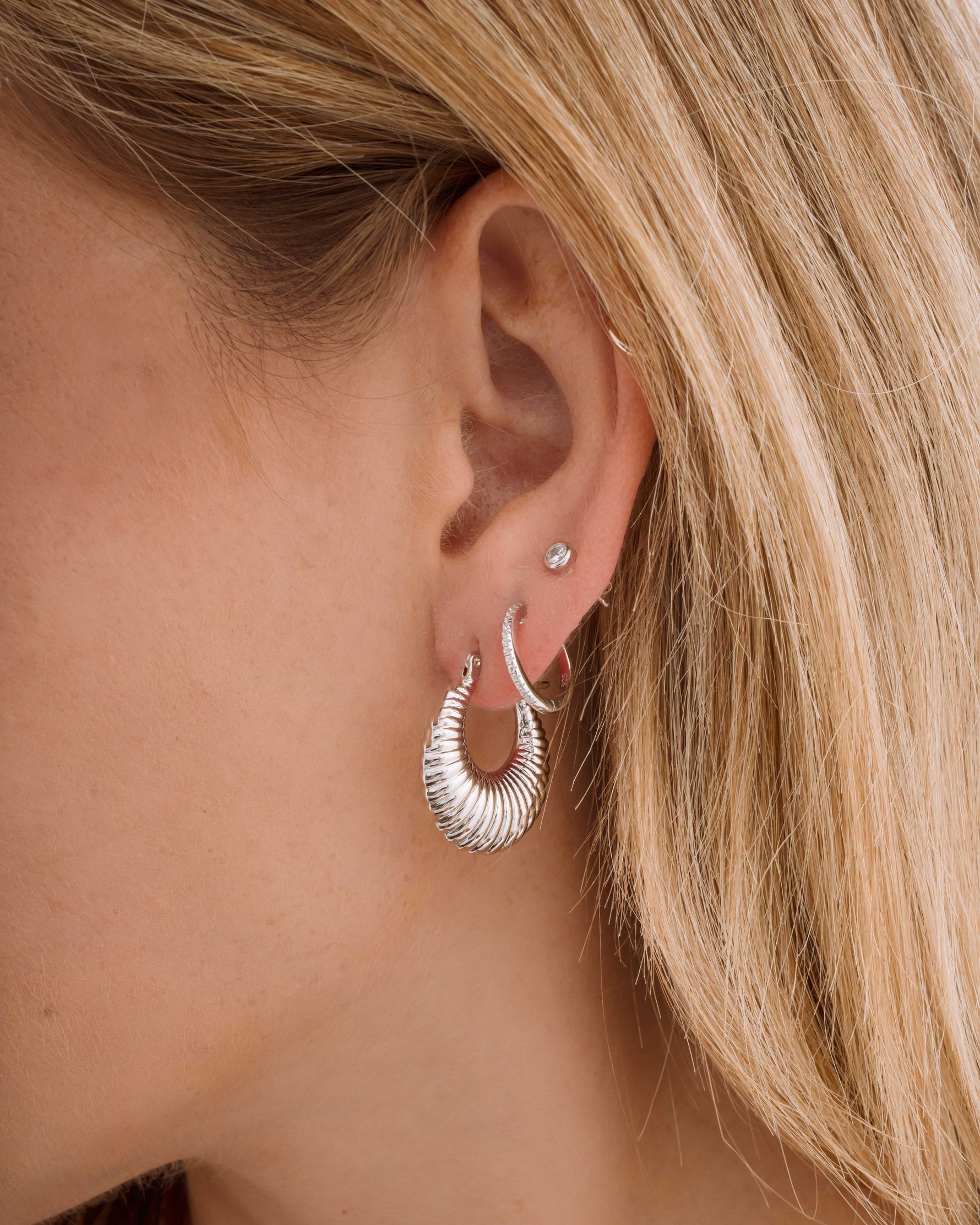 Close-up of a model wearing silver thick hoop earrings, diamond huggies, and a bezel stud earring