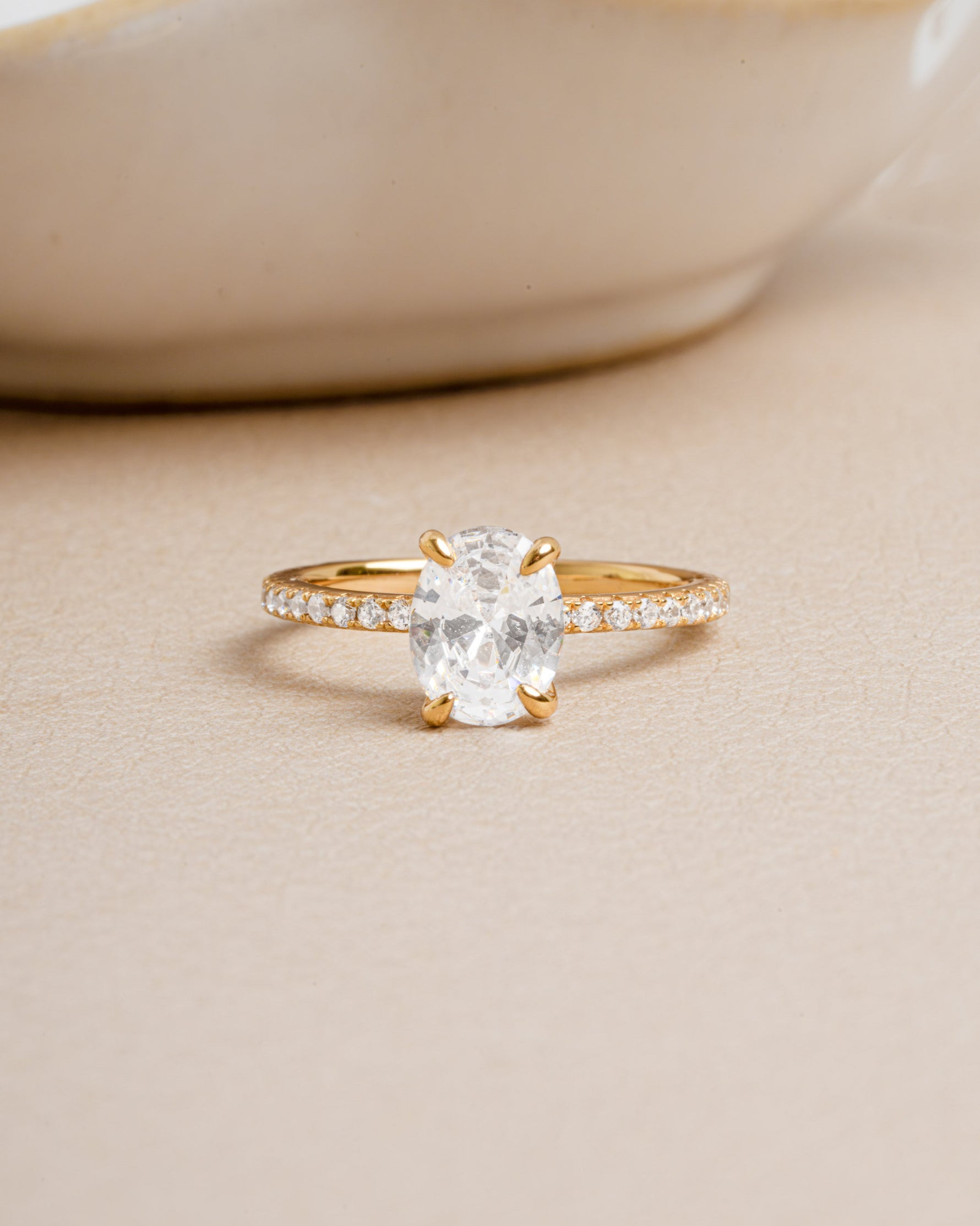Gold ring with a large oval cut diamond and pave diamond band set against a beige background.