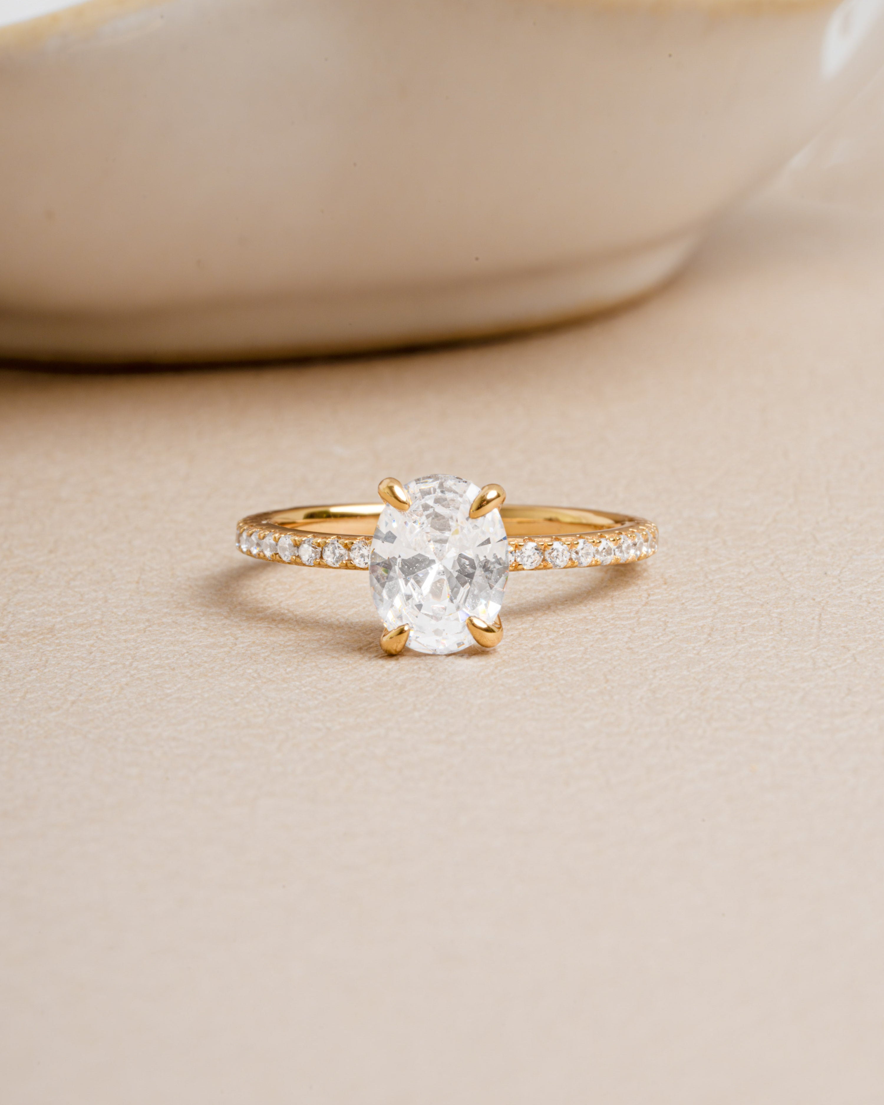 Gold ring with a large oval cut diamond and pave diamond band set against a beige background.