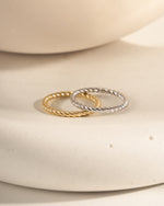 Two rings stacked on a white ceramic surface, on in 925 sterling silver with a rope ring design and one in 14k gold plating