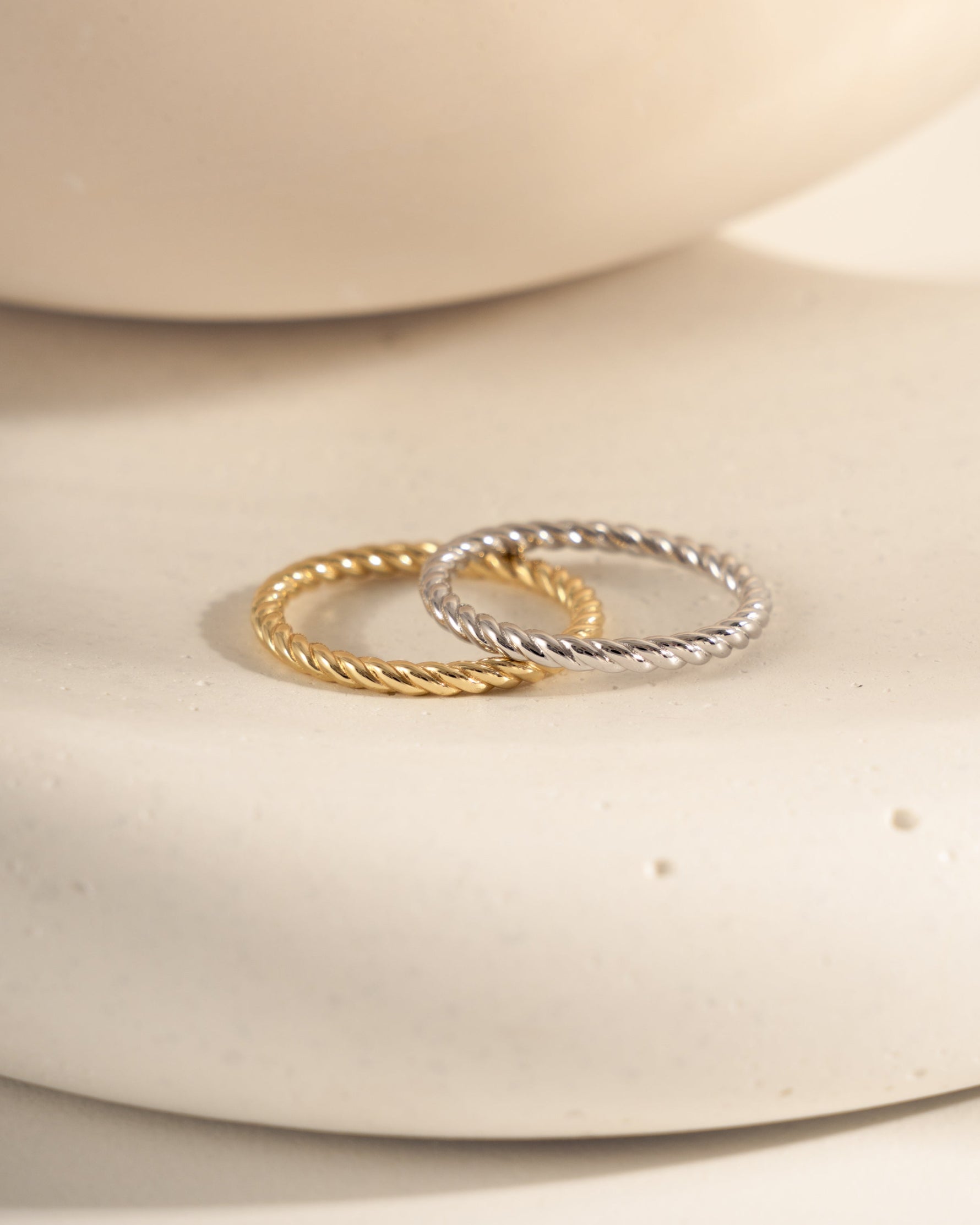 Two rings stacked on a white ceramic surface, on in 925 sterling silver with a rope ring design and one in 14k gold plating