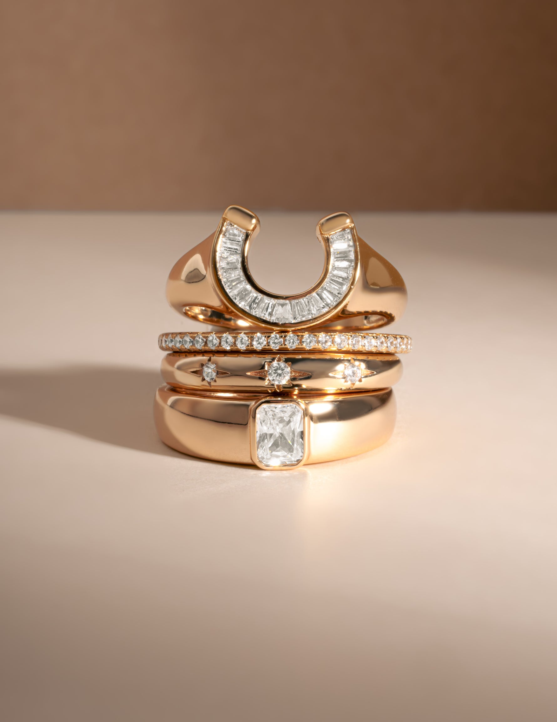 A stack of four  gold rings displayed on a warm beige surface against a matching background. The rings include a horseshoe-shaped band set with clear stones, a thin pavé band, a band with a central clear rectangular stone, and a thick band featuring a larger rectangular clear center stone.
