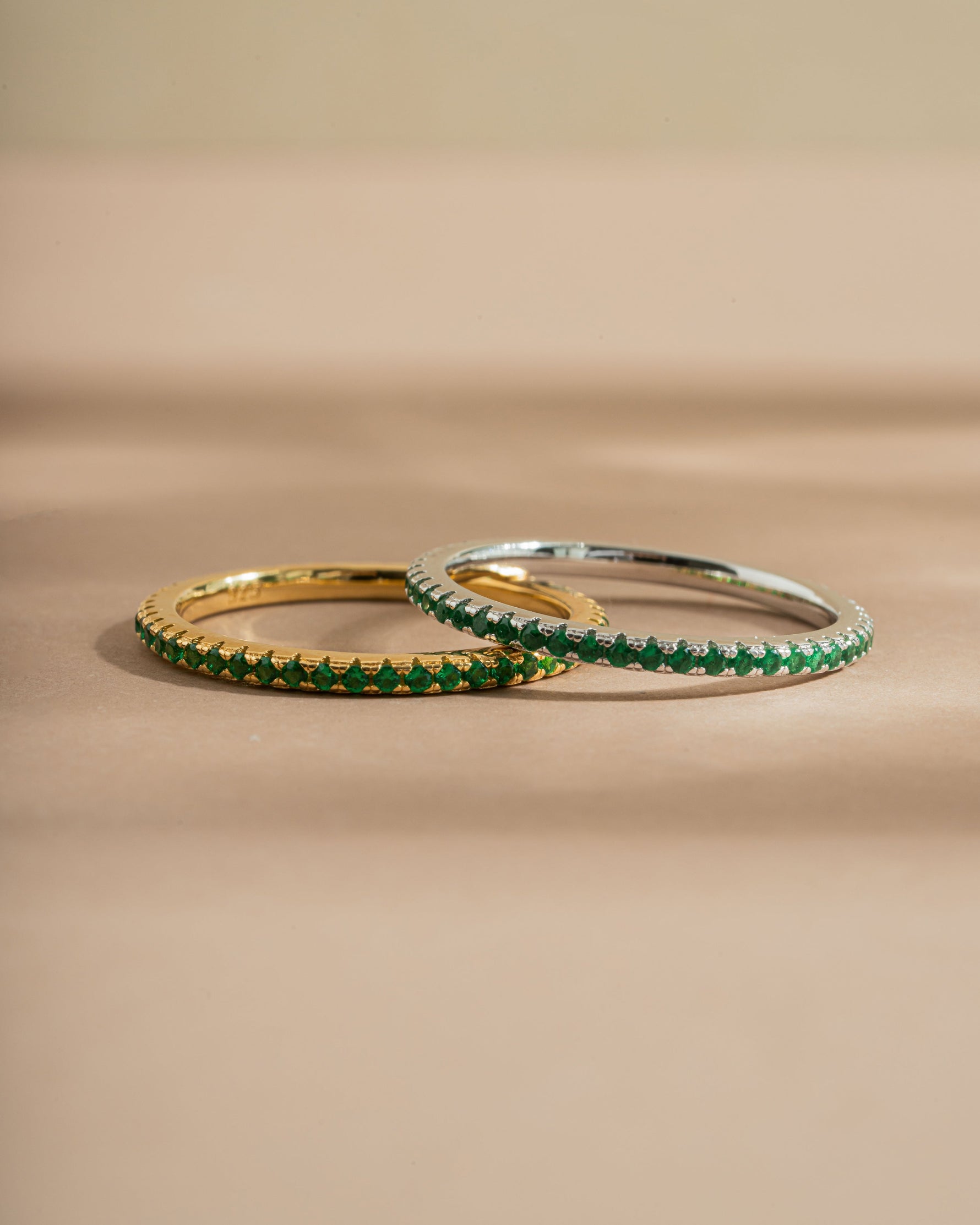 Two gold and silver eternity rings with emerald stones on a beige background