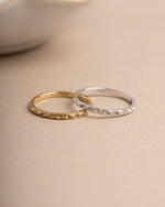 Two dainty rings on a beige surface, one gold minimalist ring and one sterling silver stacking ring