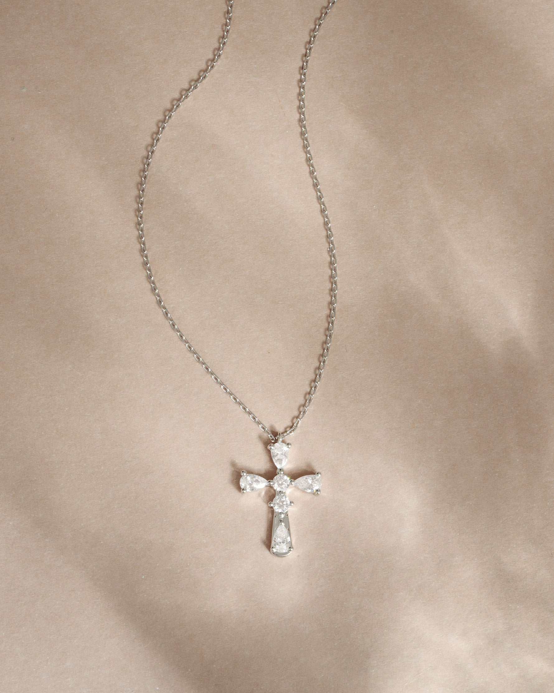 Silver chain necklace with pear cut diamonds with a cross pendant on a beige fabric background
