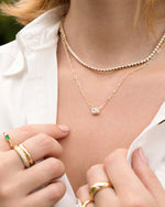 Close-up of a model wearing gold jewelry including a. diamond tennis necklace, a baguette paperclip chain necklace, and gold cz rings