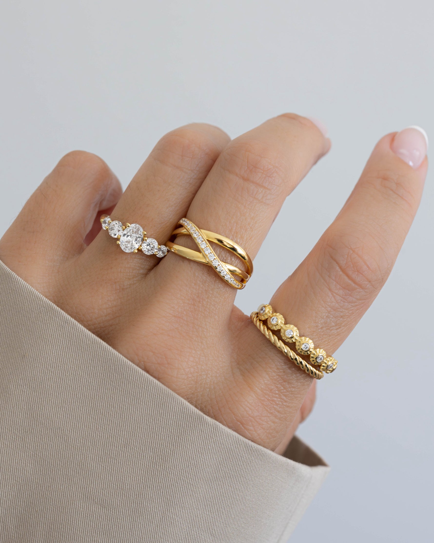 model wearing three gold rings with diamond accents on a neutral background, one ring with bezeled diamonds, a rope ring, a oval diamond ring, and a double band x ring with diamond accents