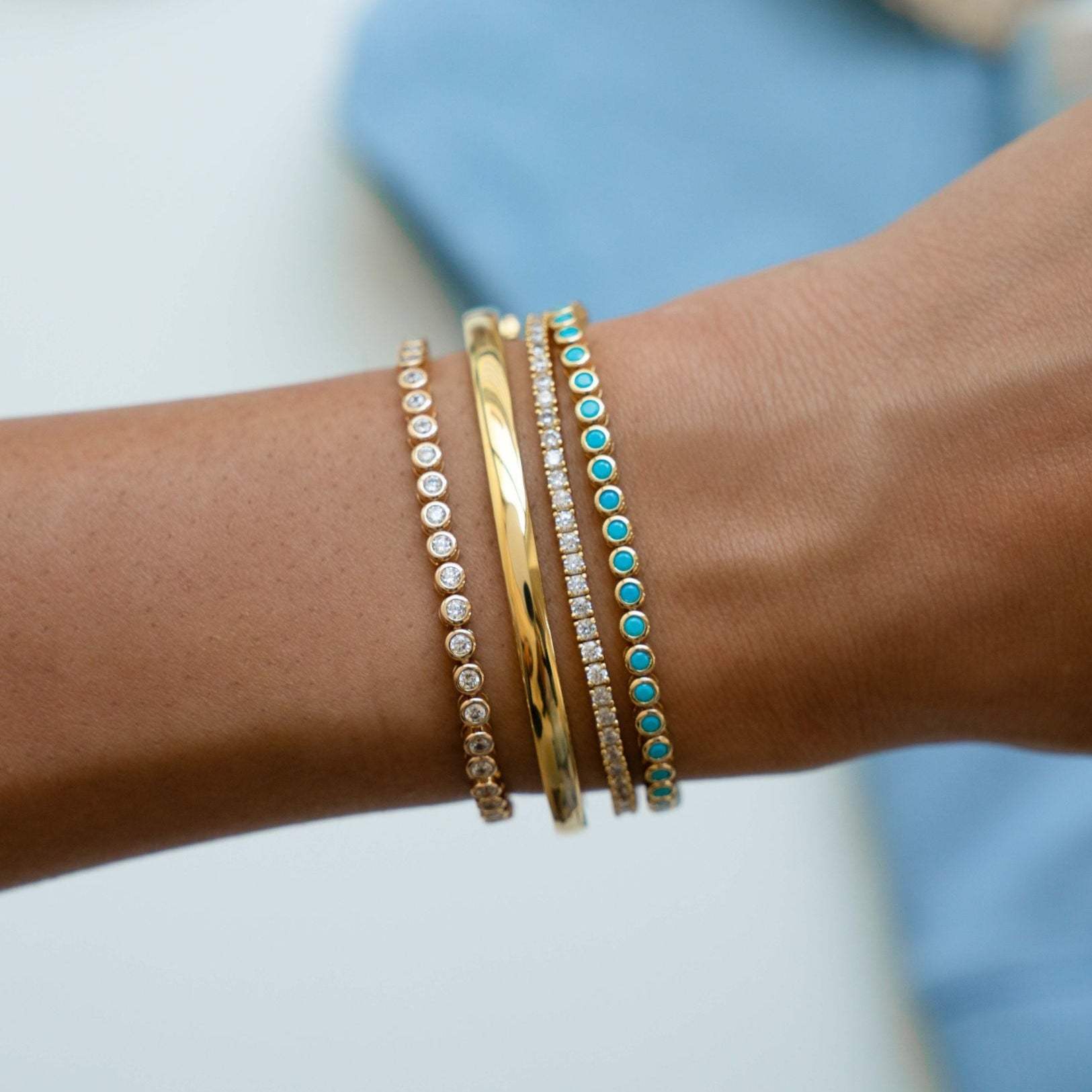 Close-up of a wrist wearing multiple Sami Jewels gold tennis bracelets bracelets, including a turquoise bezel tennis bracelet and a 14k gold plated bangle bracelet with a blurred background