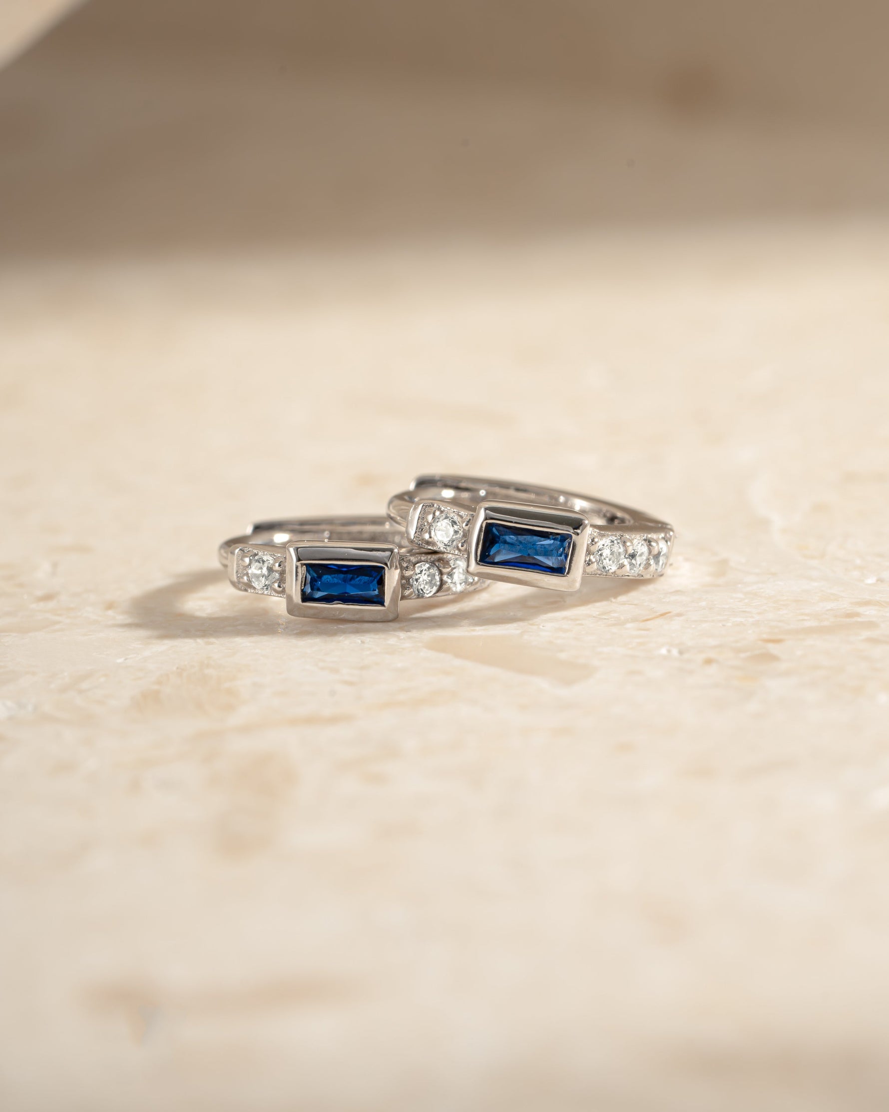 Close-up of Sami Jewels sapphire baguette huggie earrings in sterling silver, showcasing their sparkling blue sapphires and sleek design.