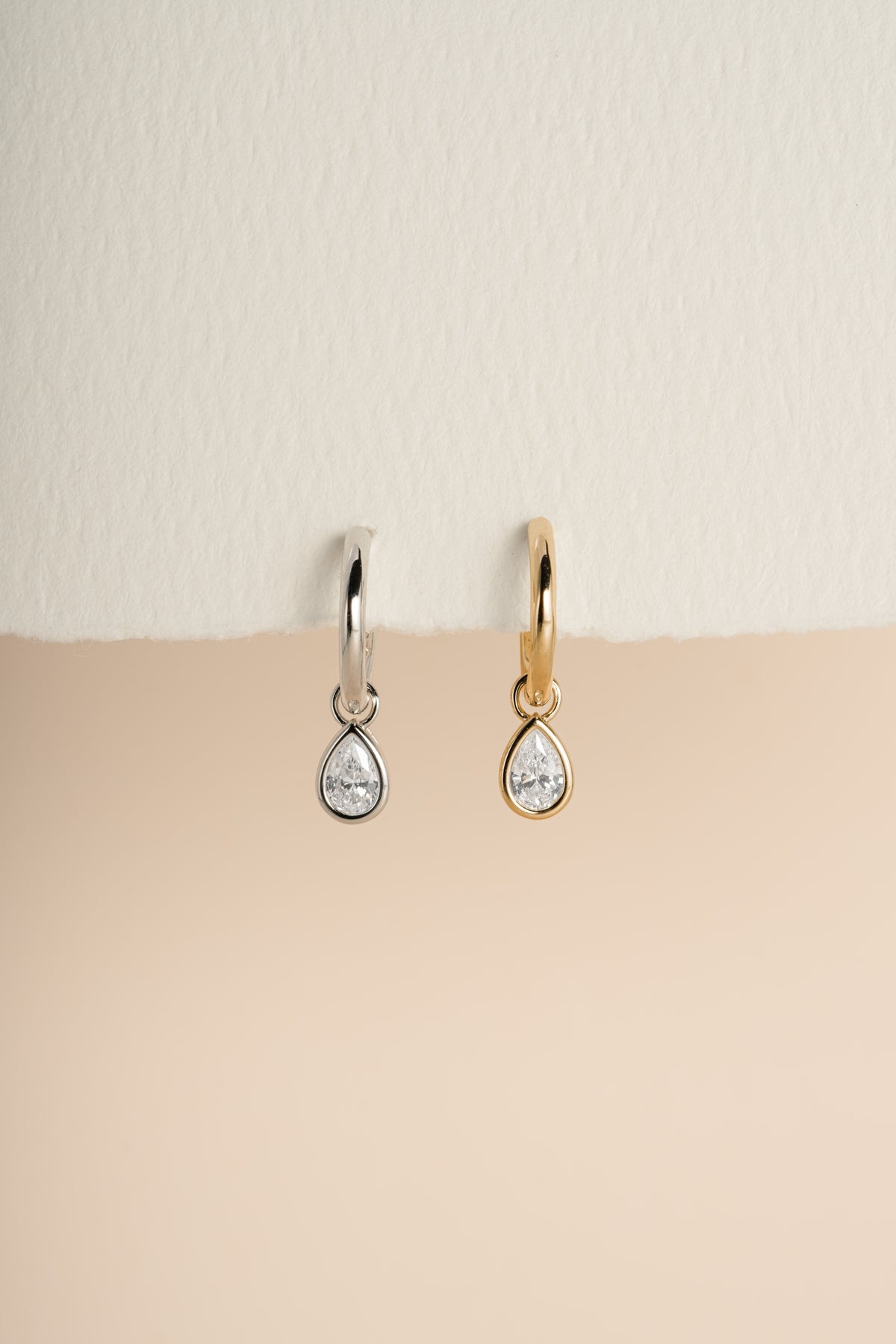 Gold and silver pear CZ charm earrings side by side against a warm neutral background.