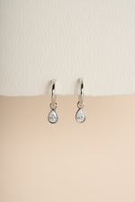 Pair of silver pear CZ charm earrings hanging against a warm neutral background.