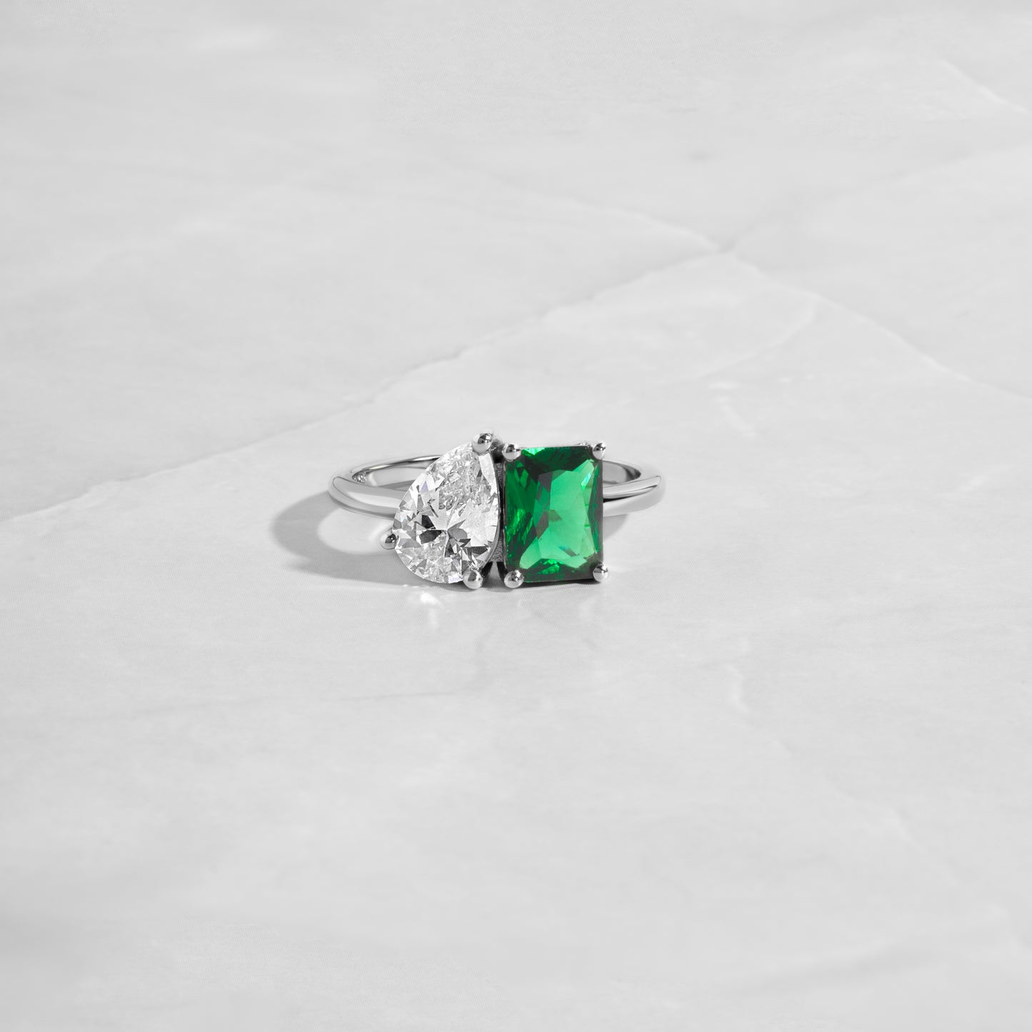 flat lay of a two stone ring in silver, showcasing the emerald cut and 
 teardrop shape diamonds, making it a standout dainty ring. 