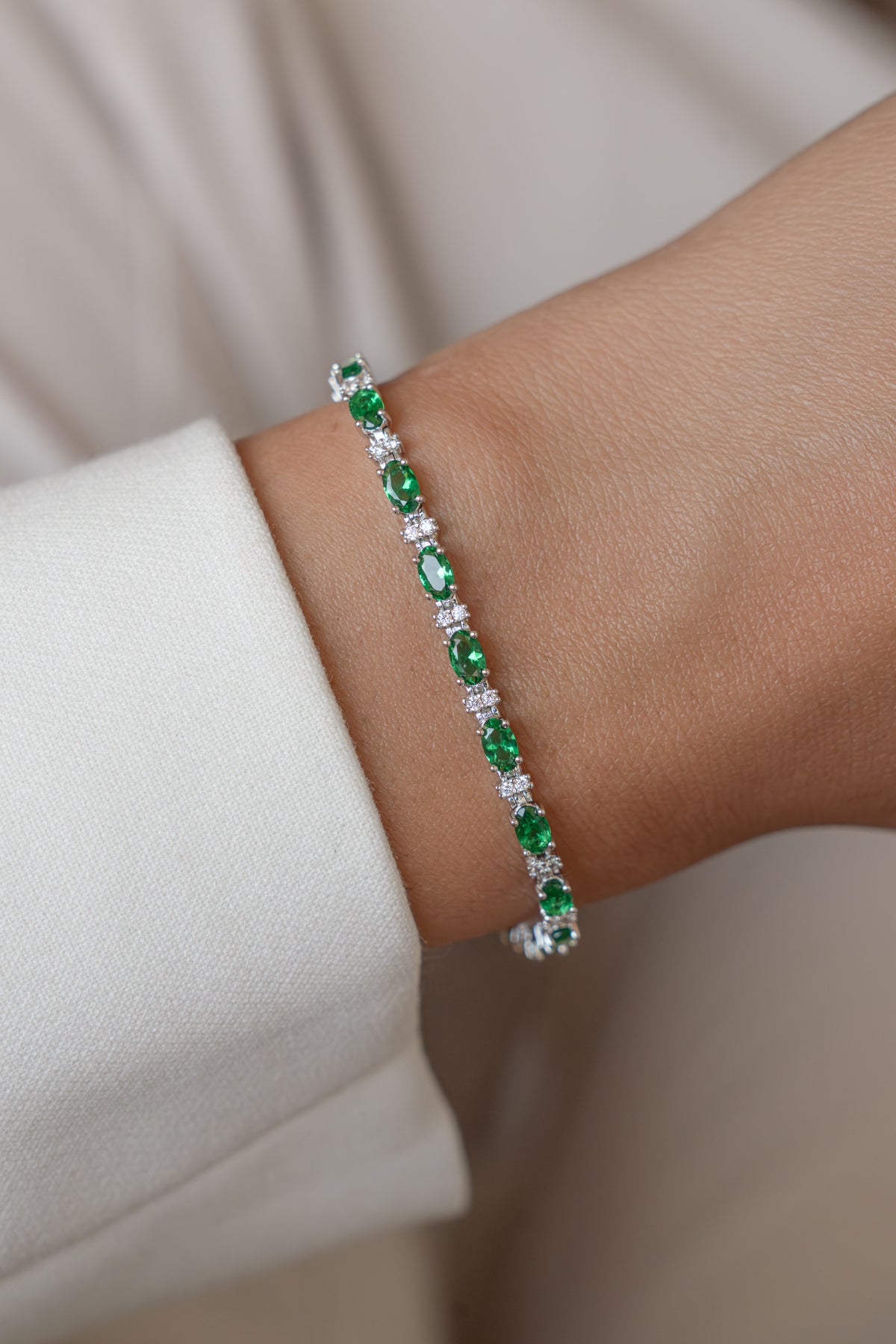 A modeled on wrist silver tennis bracelet with oval emerald and round white gemstones. 