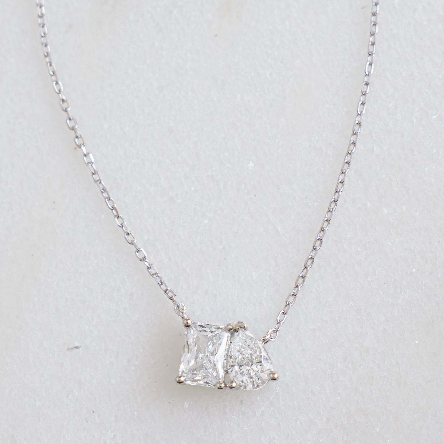 A flat lay of dainty silver necklace, featuring an emerald cut stone set against a pear shaped stone symbolizing two lovers known as a toi et moi design