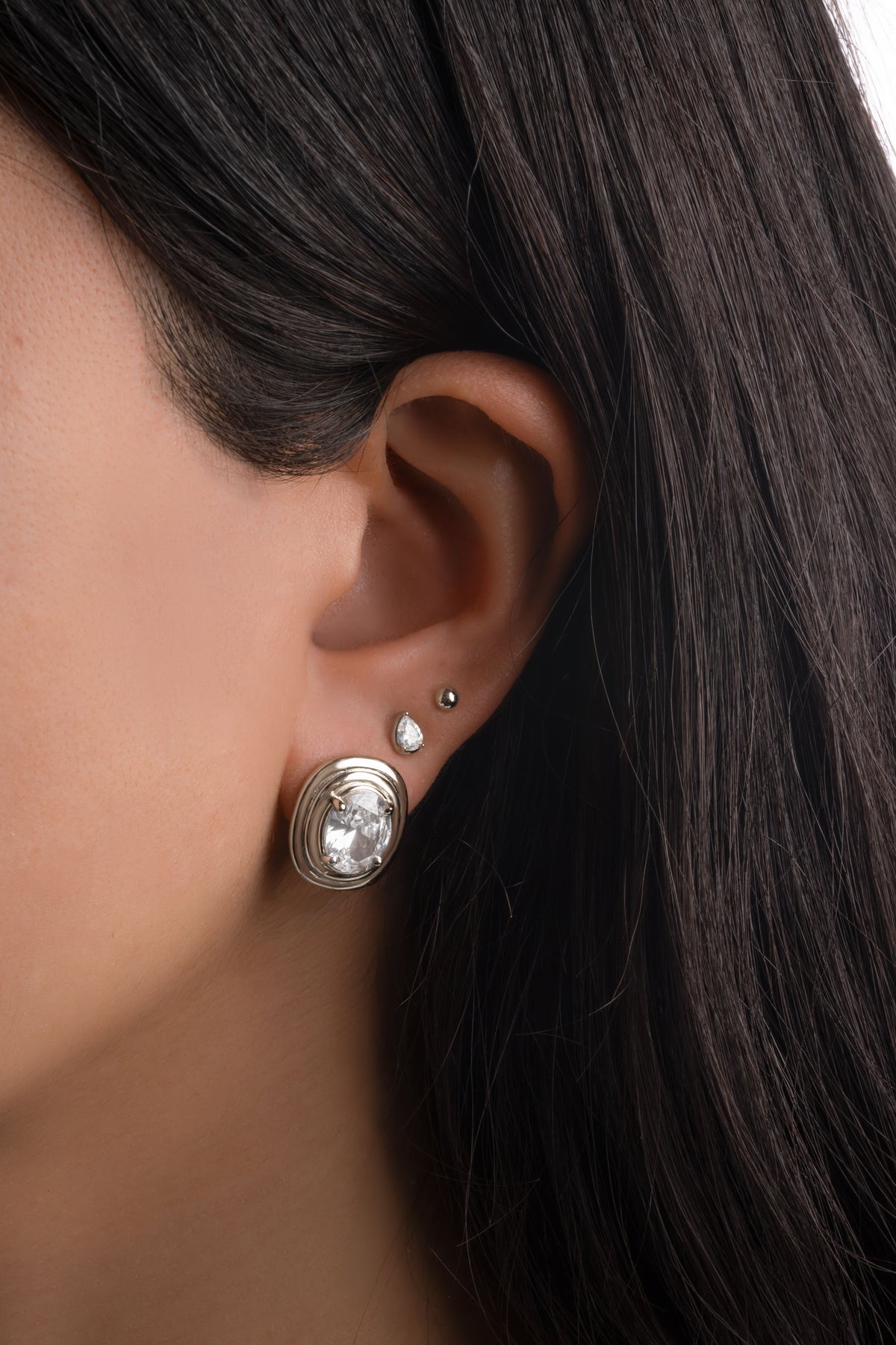 Silver oval bezel statement stud on a model's ear, styled with a pear-shaped stud and a small ball stud.
