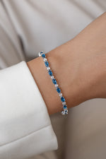 An oval-cut sapphire tennis bracelet with alternating white diamonds with a silver finish modeled on a wrist.