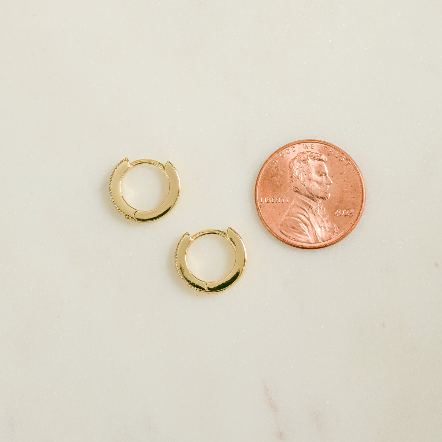 Close up flat-lay image of minimalist earrings. The gold baby huggie earrings are being compared to the size of a penny. 