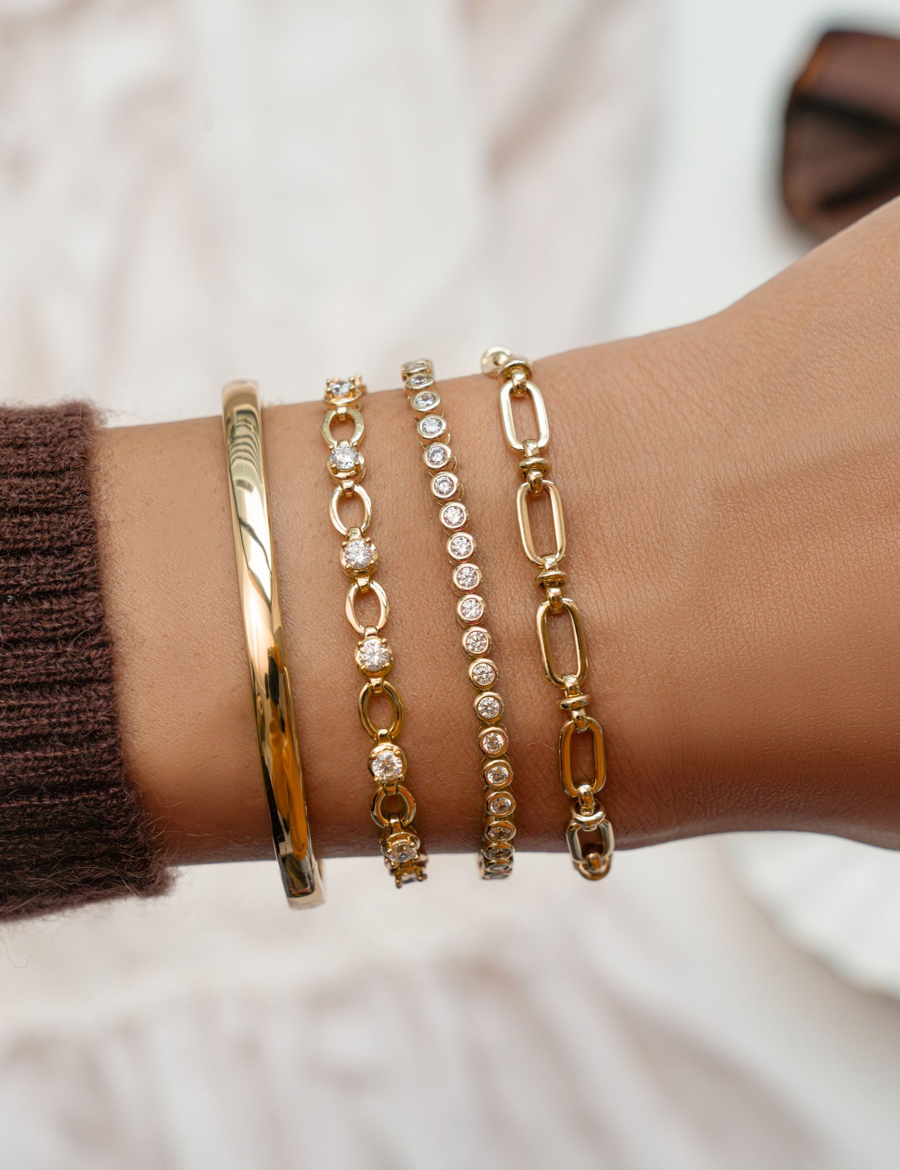 Solid gold bangle stacked with tennis bracelets and a chunky chain link bracelet on wrist with blurred background