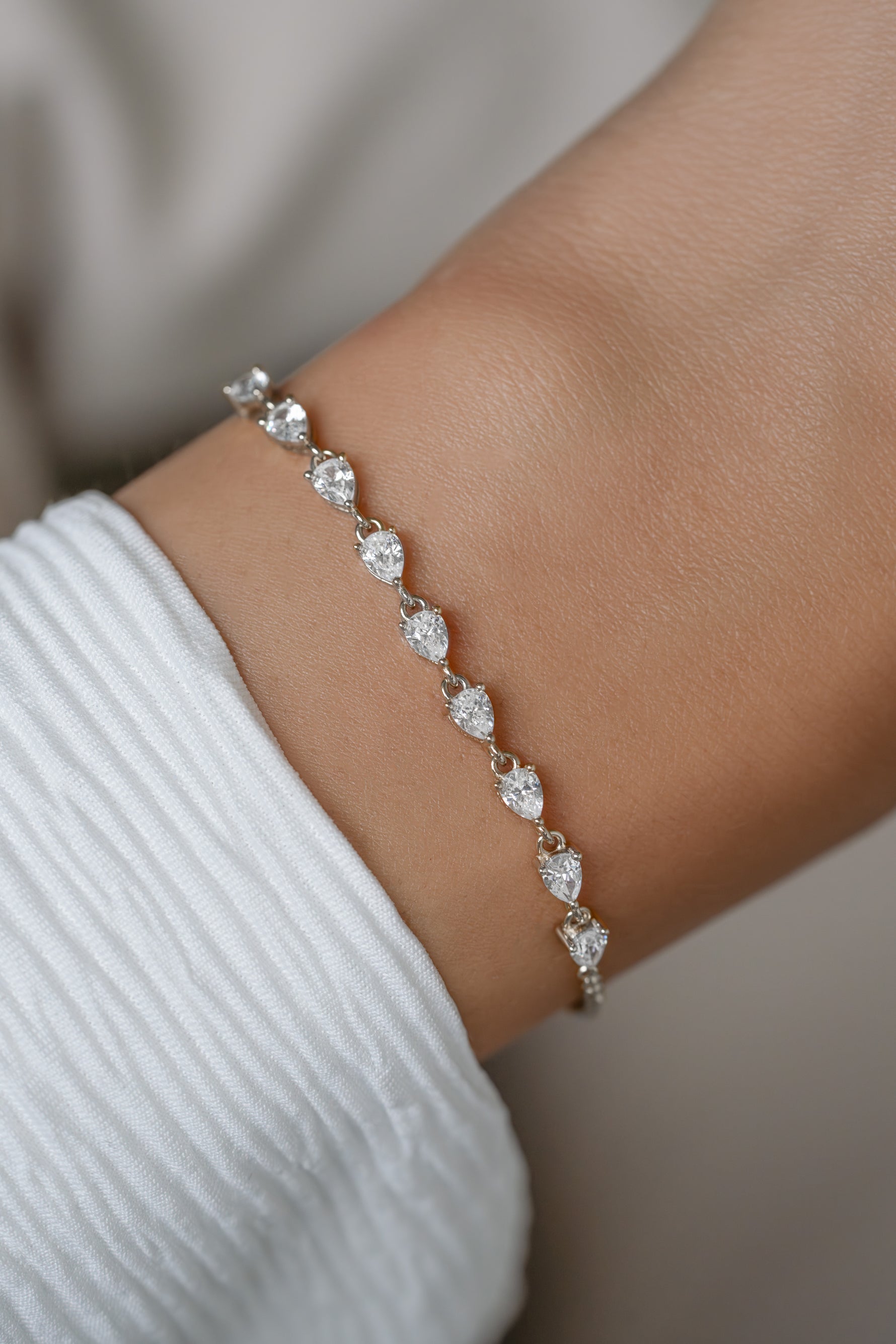 A delicate tennis bracelet with clear pear-cut gemstones in a silver-toned setting, shown worn on a person's wrist over a white ribbed sleeve.