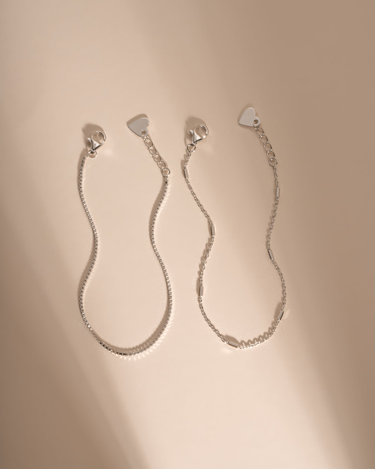 Two silver bracelets laid side by side, showing their lobster clasps, extenders and heart-shaped tags.