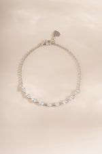A delicate silver chain bracelet with a series of cubic zirconia stones shaped in pear or tear drop set along the lower curve, featuring a small heart-shaped charm at the clasp, displayed on a soft beige background.
