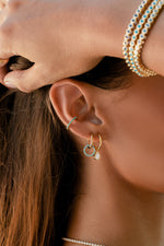 Close-up of a gold pear CZ charm earring styled with turquoise-colored earrings and bracelets on a model.